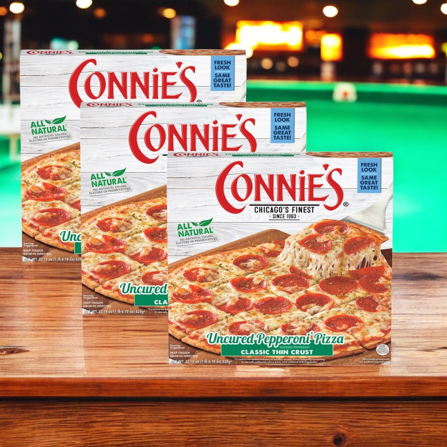 Displayed on a wooden surface with a blurred background, three packs of Connie's Pizza Classic Thin Crust Uncured Pepperoni offer a delicious and convenient meal option without compromising on flavor.