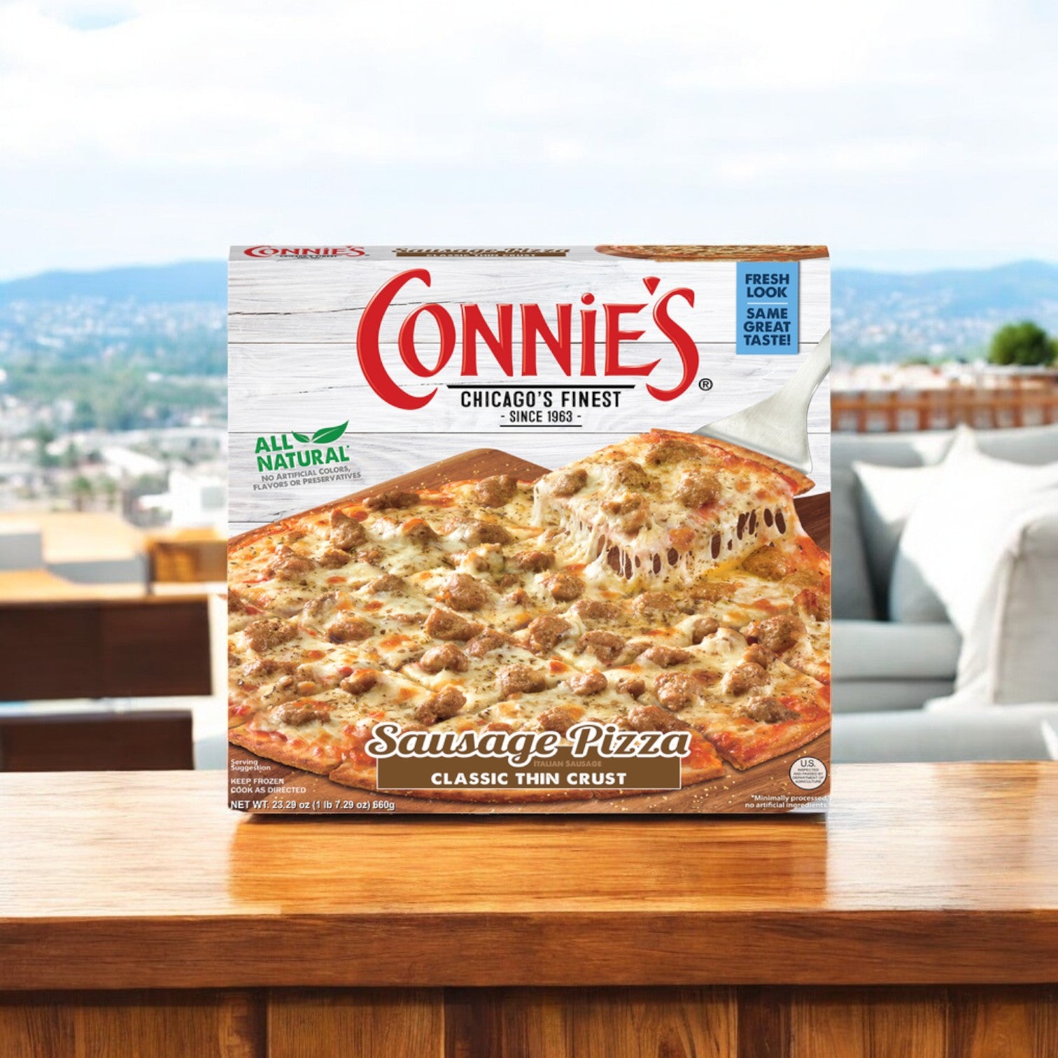 A box of Connie's Pizza Sausage, Classic Thin Crust, weighing 23.29 oz., is placed on a wooden table outdoors, complemented by a scenic view in the background.