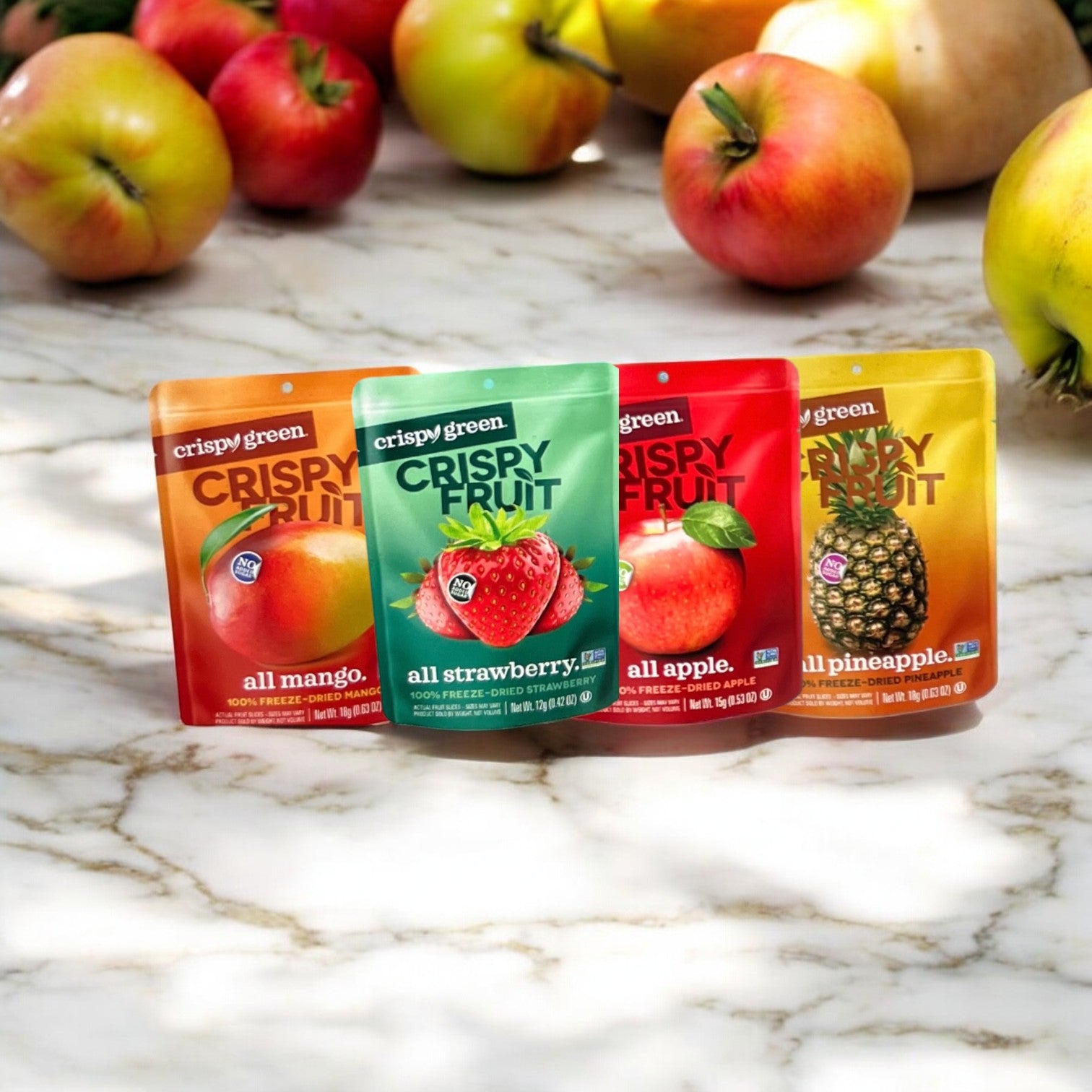 A variety of Easy Lunches' Crispy Fruit Best Selling Variety Pack, featuring all-mango, all-strawberry, all-apple, and all-pineapple freeze-dried fruit snacks, are displayed on a marble surface with fresh apples surrounding them to complement their crispy texture and natural sweetness.