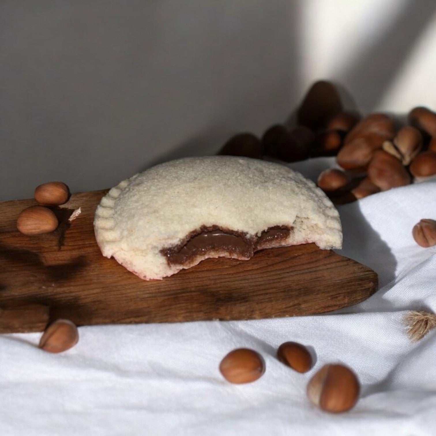 A partially eaten Uncrustables Chocolate Flavored Hazelnut Spread Sandwich by Uncrustable Smuckers, sitting on a wooden board with unshelled hazelnuts scattered around.