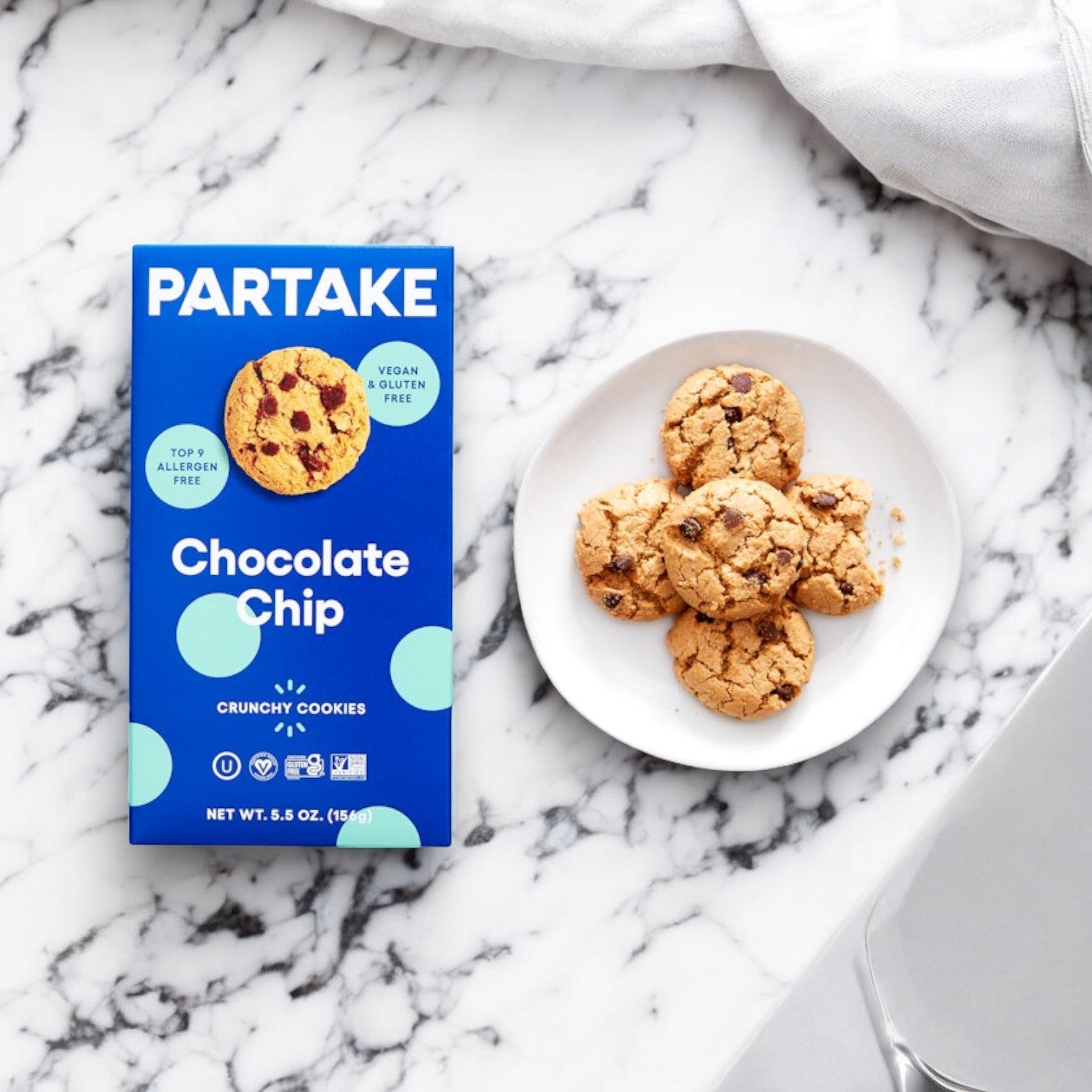 A box of Partake Gluten Free Vegan Chocolate Chip Cookies- 1 Box placed on a marble surface, next to a white plate containing five cookies, reflects Partake's commitment to inclusivity and access for all cookie lovers.