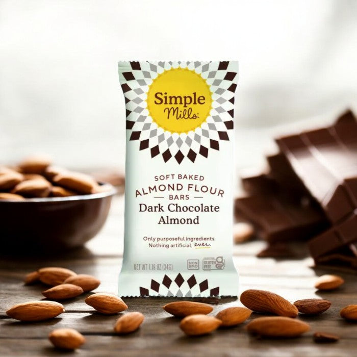 A package of Simple Mills Soft Baked Almond Flour Bars in Dark Chocolate Almond flavor, surrounded by almonds and pieces of chocolate.