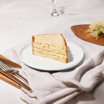 A slice of layered cake on a white plate with a fork and knife on the left, placed on a light-colored cloth. In the background, there is a glass and a wooden board with decorations, possibly set for enjoying some Taylor Farms' Tuna Salad Sandwich.