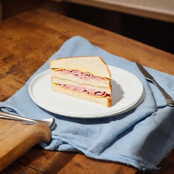 A Taylor Farms Ham and Cheddar Sandwich, made with white loaf bread and featuring smoked ham and mild cheddar cheese, is cut diagonally and placed on a white plate atop a light blue cloth on a wooden table, next to a butter knife and fork.