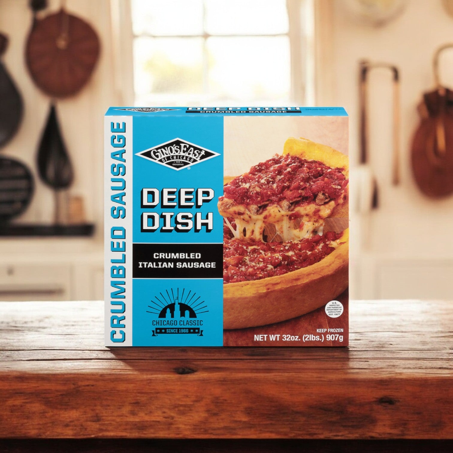 A box of Gino's East Crumbled Sausage Deep Dish Pizza, 32 oz., sits on a wooden counter with a kitchen background. The packaging highlights the product and nutritional information, emphasizing the use of premium cheeses.