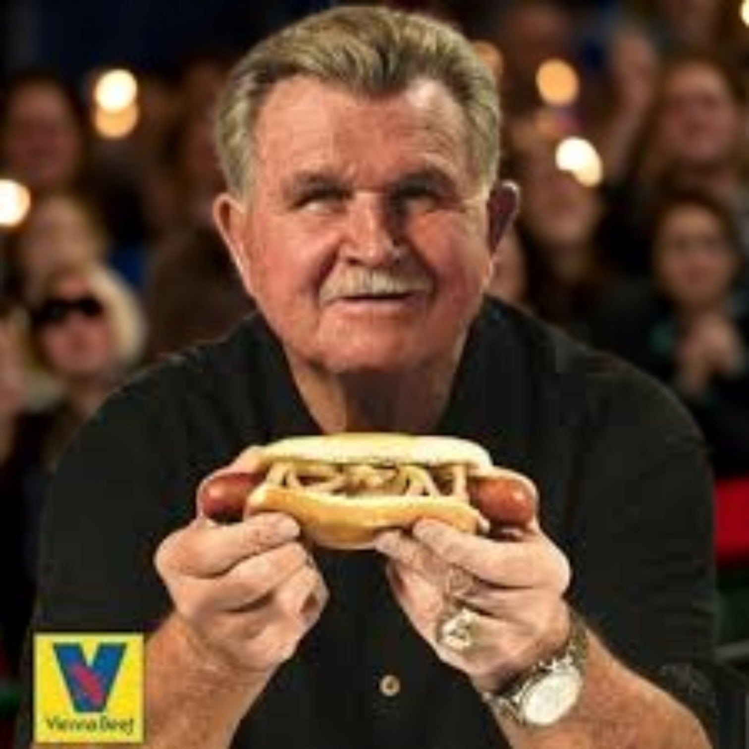 A man holds a Ditka Hot Beef Polish Sausage from Vienna Beef, loaded with onions and mustard, in front of a crowd. The Vienna Beef logo in the corner pays tribute to Mike Ditka's iconic taste for Chicago's finest.