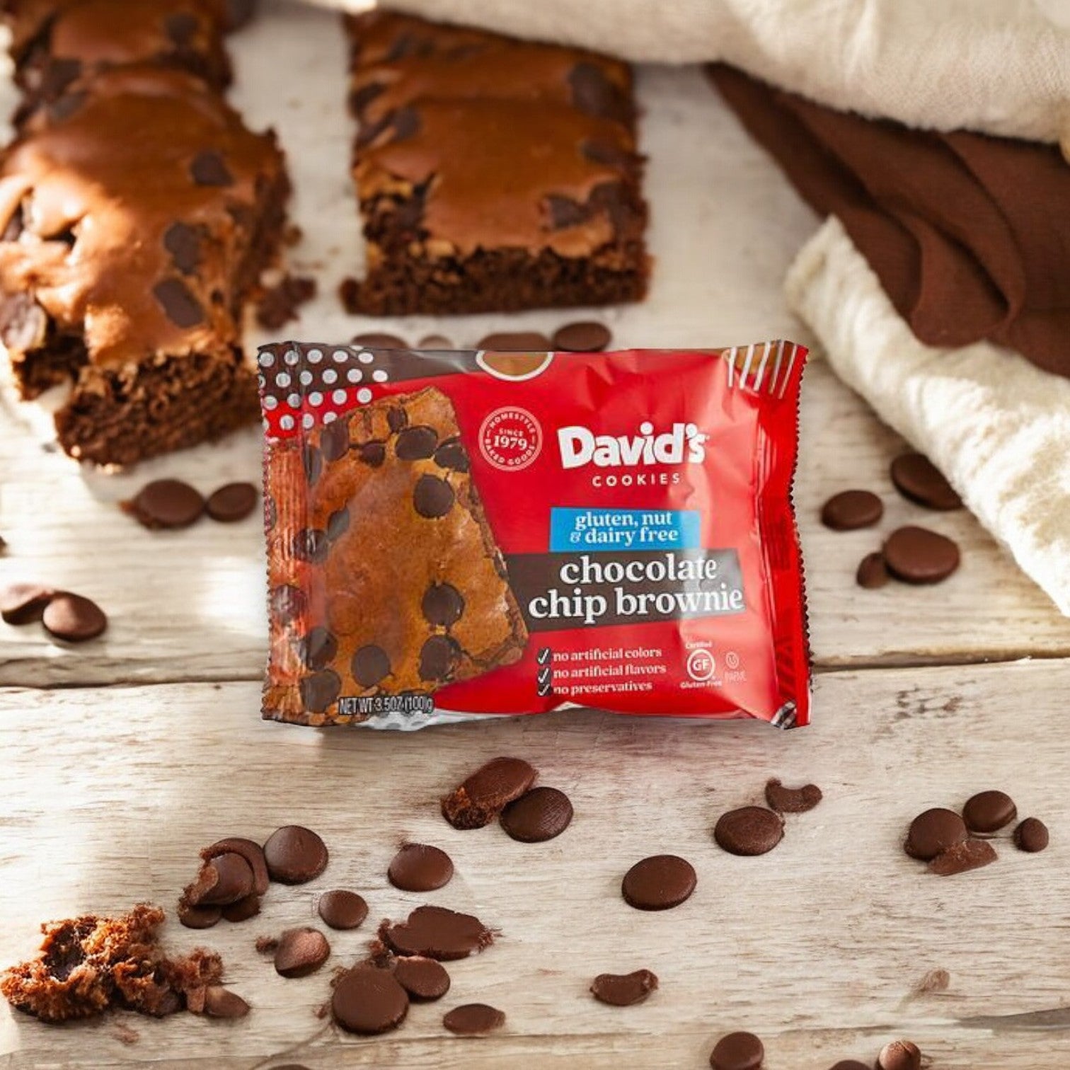 A packaged David's Cookies Gluten-Free Individually Wrapped Chocolate Chip Brownie 3.5 oz - 1 Count sits on a wooden surface surrounded by chocolate chips and cloth napkins. The brownie bar is partially eaten, revealing its rich, tempting interior.