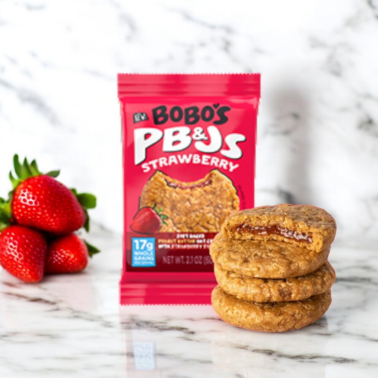 A package of Bobo's PB&Js Peanut Butter Oat Crust With Strawberry Filling, 2.1 oz - 1 Count stands upright on a marble surface beside fresh strawberries and a stack of three vegan, whole grain oats snack bars, one with a bite taken out, revealing the strawberry filling.