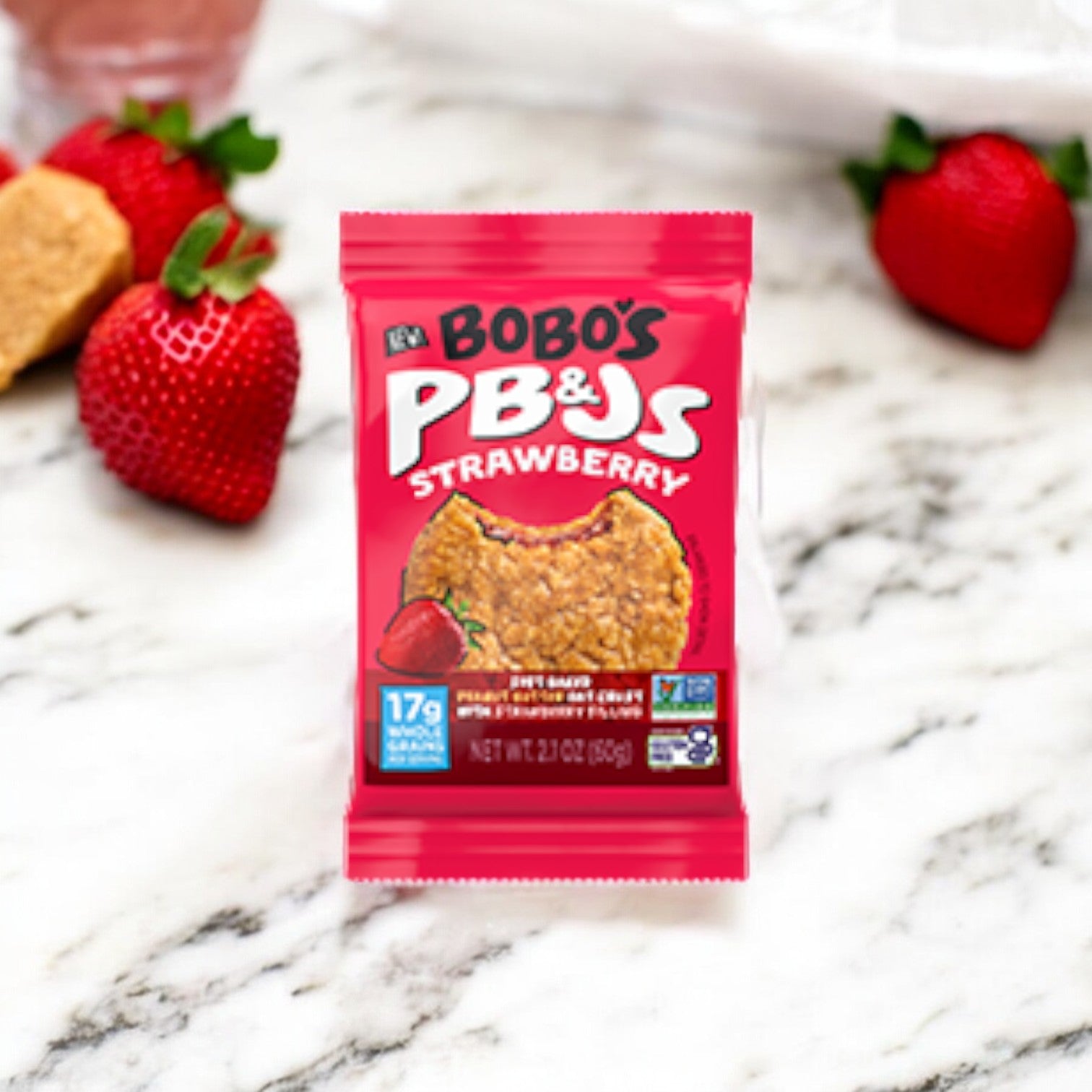 Image of Bobo's PB&Js Peanut Butter Oat Crust With Strawberry Filling on a marble surface surrounded by fresh strawberries and oat pieces. The packaging is red with text indicating high protein content and other nutritional information, showcasing its vegan and gluten-free whole grain oats.