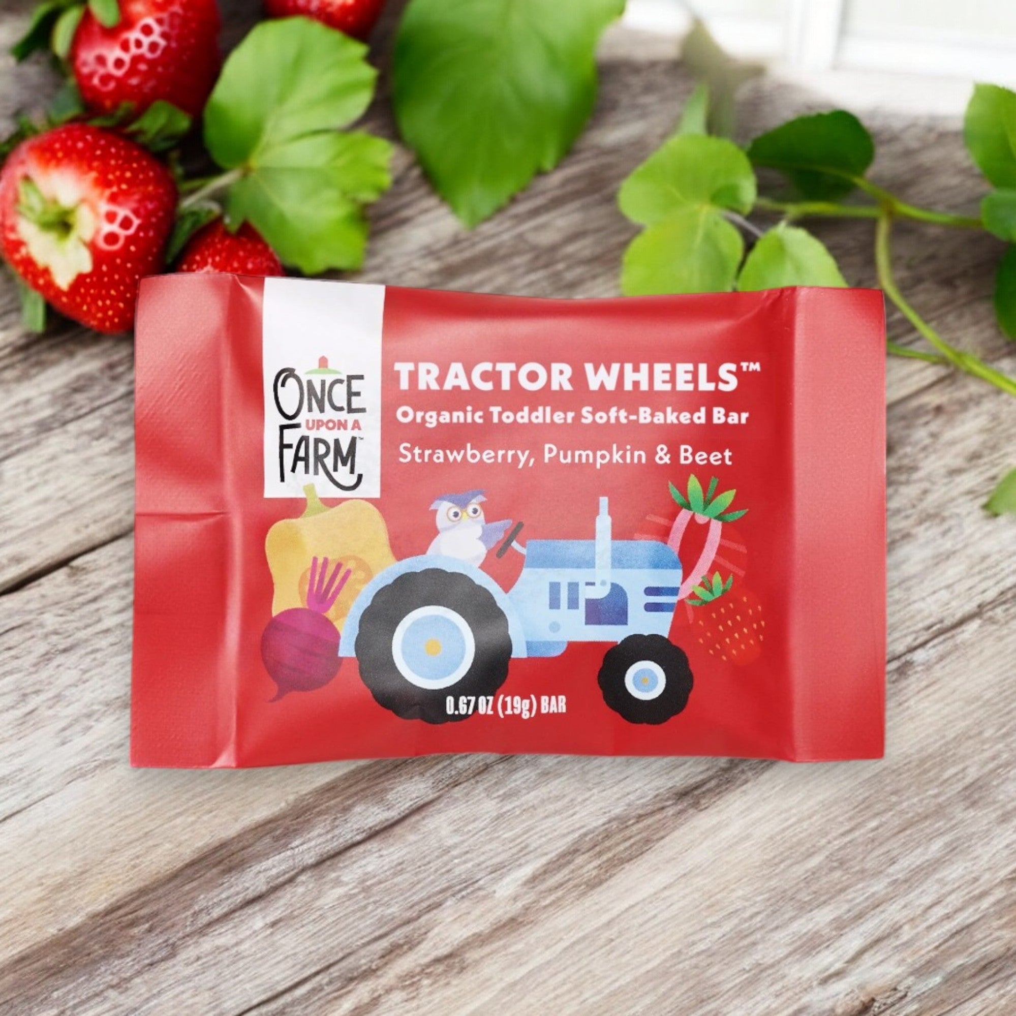 A red packet labeled "Once Upon a Farm Organic Tractor Wheels Toddler Bar, Strawberry, Pumpkin & Beet" is placed on a wooden surface next to fresh strawberries and green leaves, featuring whole grain oats as the perfect organic snack for toddlers.