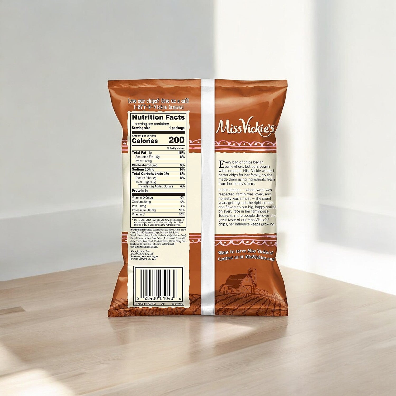 A 1.375 oz bag of Miss Vickie's Smokehouse BBQ Flavored Kettle Cooked Potato Chips, featuring the nutritional information, ingredients, and branding prominently displayed on the back of the packaging. The chips bag is positioned on a light wooden surface, illuminated by soft natural light.