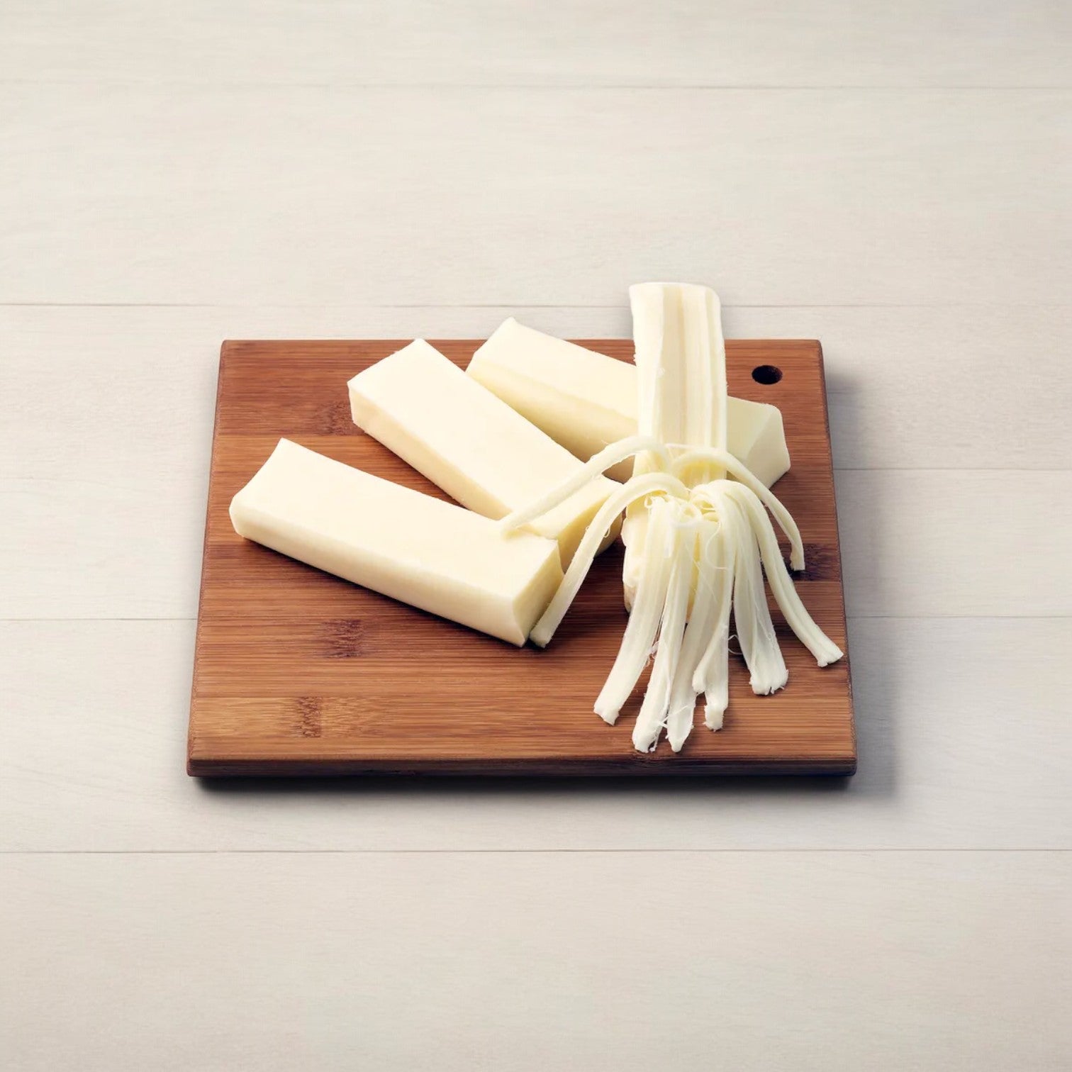 Three sturdy sticks and one stringy piece of Frigo® Cheese Heads® Original Mozzarella String Cheese Snacks, made from low-moisture part-skim mozzarella, are elegantly arranged on a wooden cutting board placed on a light-colored surface. This high-protein snack from Frigo is perfect for any occasion.