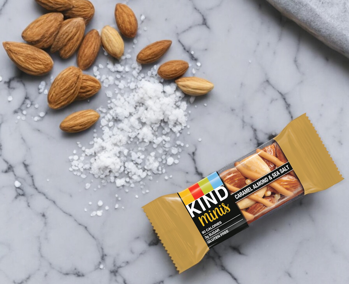 A Kind Minis Caramel Almond & Sea Salt bar from the 10-count pack by Kind is placed on a marble surface, surrounded by scattered almonds and sea salt, offering a delicious and healthy snack.