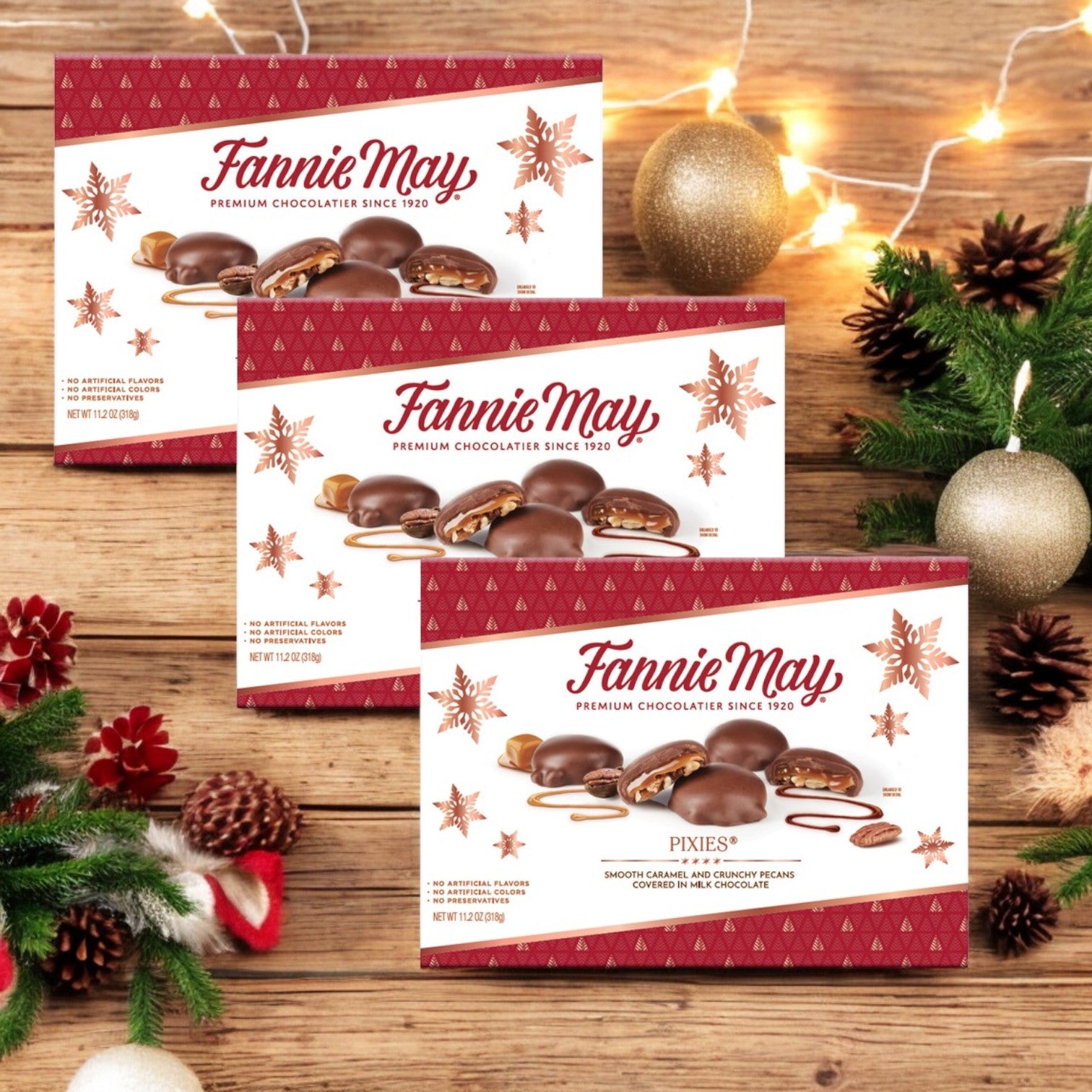 Three charming boxes of "Fannie May Chocolates, Pixies, 3 Count" rest on a wooden table adorned with shimmering Christmas decorations and lights, their milk chocolate caramel treats promising a delightful holiday indulgence.