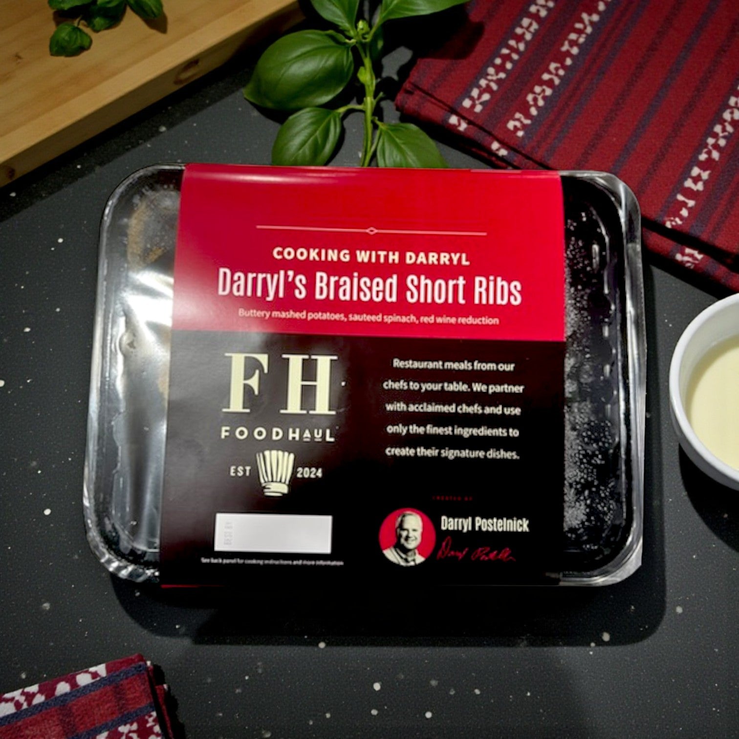 A FoodHaul Frozen Meal—Darryl's Braised Short Ribs by Darryl Postelnick—rests on a kitchen counter beside a red napkin, small bowl, and cutting board.