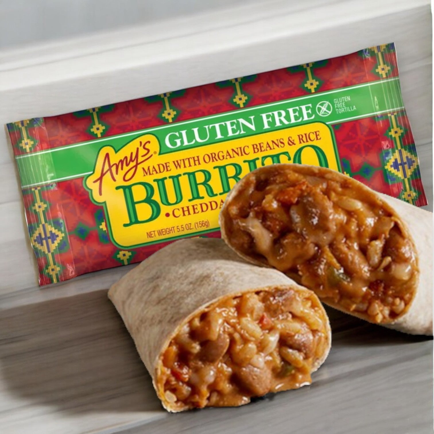 A packaged Amy's Gluten Free Bean & Cheese Frozen Burrito sits behind a split burrito revealing its rice, beans, and cheddar cheese filling on a marble surface, showcasing the delicious combination of organic ingredients from Amy's.