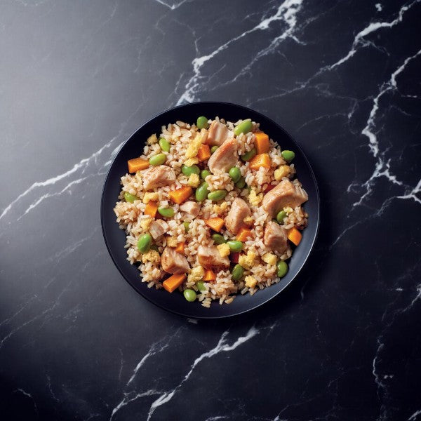 A black plate brimming with chicken fried rice, tender chicken pieces, edamame, and diced carrots, set against a dark marble background, highlights the vibrant flavors of Healthy Choice Simply Steamers Chicken Fried Rice (10 oz.) and their innovative steam-in-bag technology.