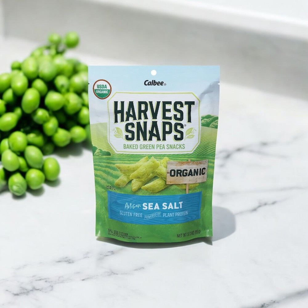 A 3 oz bag of Harvest Snaps Organic Artisan Sea Salt Baked Green Pea Snacks is placed on a marble surface next to a bunch of fresh green peas. The packaging highlights that these gluten-free snacks are packed with plant protein.