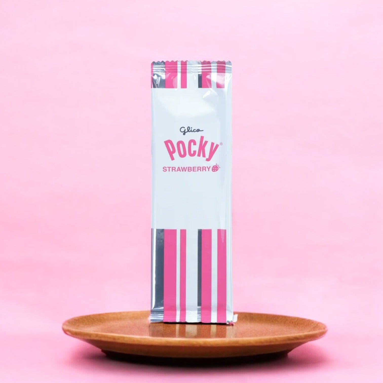 Set against a pink backdrop, this delightful package of Pocky Strawberry Cream Covered Biscuit Sticks, weighing 1.41 oz from the brand Pocky, rests elegantly on a brown plate, ready to offer a fruity and sweet experience with each crunchy stick.