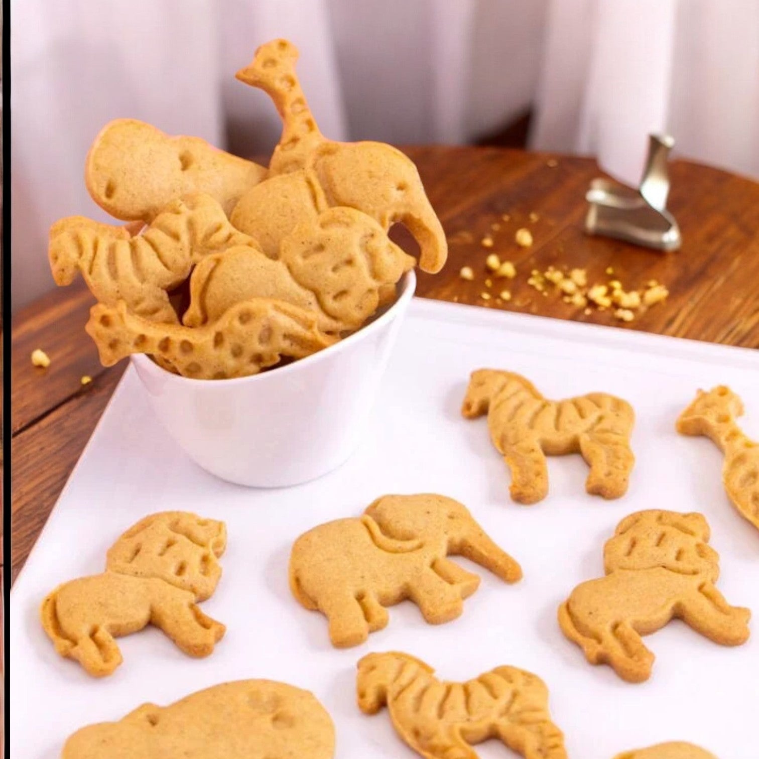 A Barnum's Original Animal Crackers 1 oz packet from Barnum's is seen spilling its delightful, calcium-rich treats into a white bowl on a wooden table. Various cookies shaped like animals, including elephants and giraffes, are scattered on a white sheet next to the bowl.