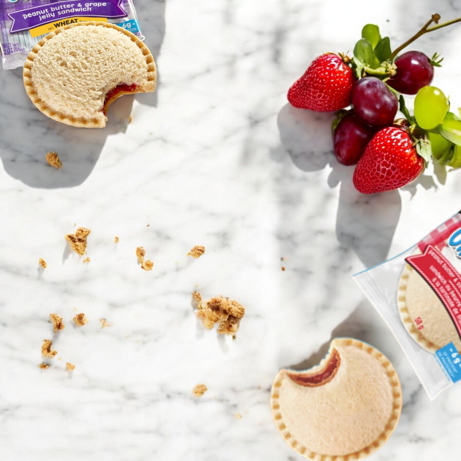 Two partially eaten Uncrustables from the Uncrustable Smuckers 10 Bundle Pack - 2 Flavors rest on a marble surface, surrounded by scattered crumbs and a small bunch of strawberries and grapes.