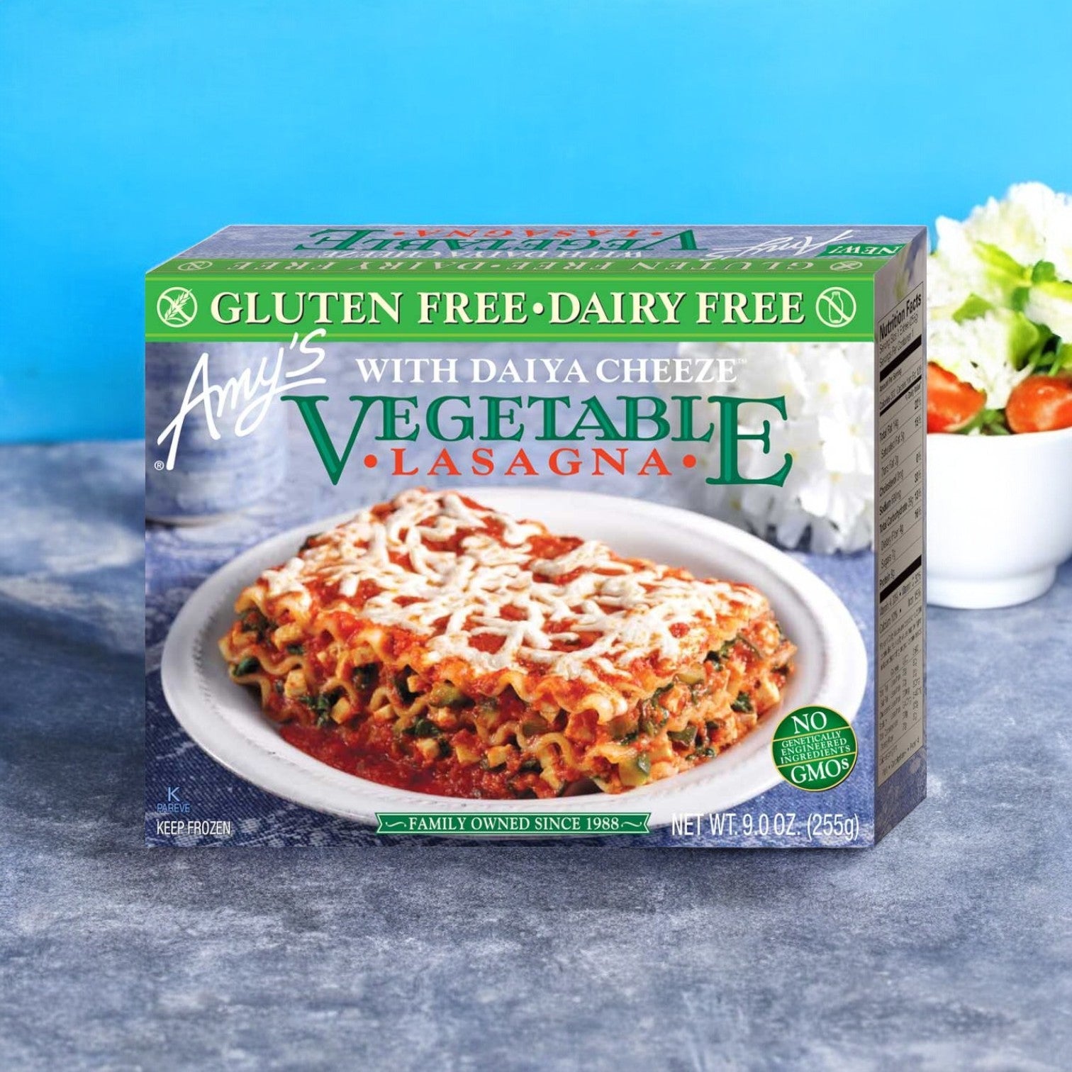 A 9 oz. box of Amy's Gluten-Free Dairy-Free Vegetable Lasagna, available in cases of 12, featuring Daiya Cheeze and made with organic ingredients, is placed on a blue surface with a salad in the background.