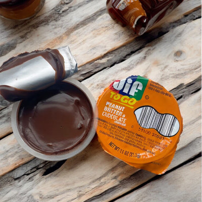 An open container of JIF To Go Spread, Peanut Butter & Chocolate Flavored, 1.1 oz with its foil lid peeled back, placed on a wooden surface. Other unopened containers of JIF are visible in the background, making it an inviting and healthy snack option.