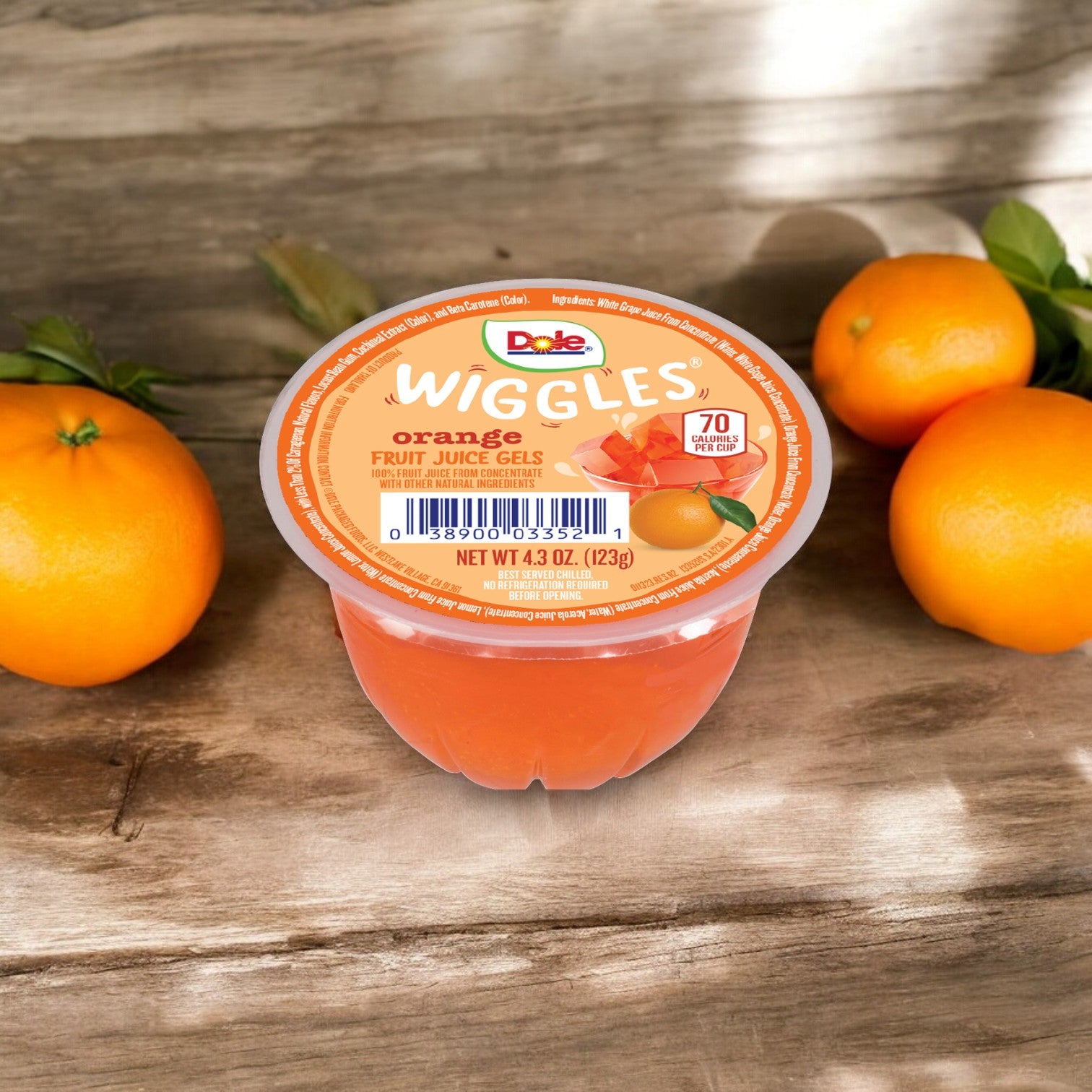 A Dole Wiggles Orange Fruit Juice Gels, 4.3 oz - 1 Count, known for its Vitamin C and no added sugar, is placed on a wooden surface with two oranges and green leaves in the background.