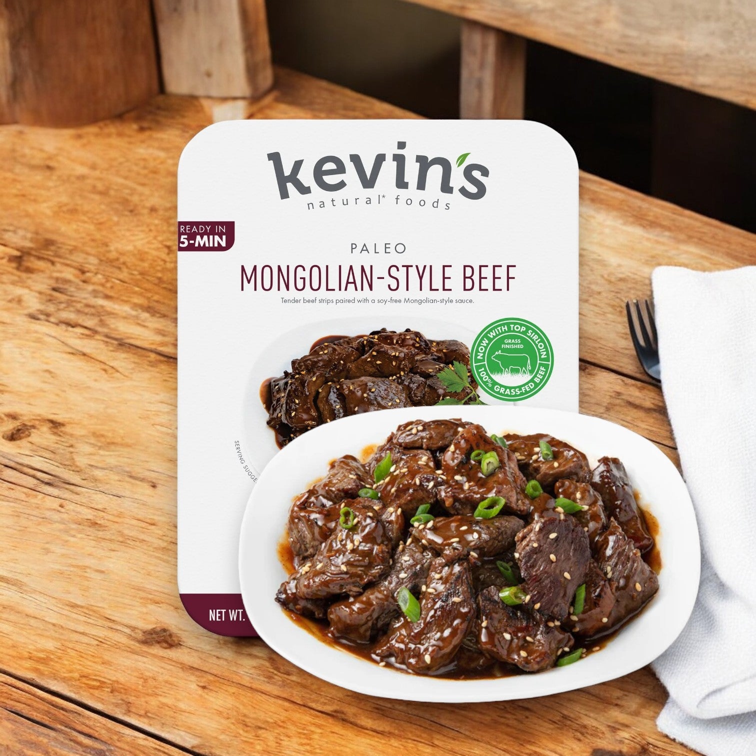 A box of Kevin's Mongolian Beef, 32 oz - 1 Count by Kevin's sits next to a plate of the prepared, gluten-free dish garnished with green onions on a wooden table with a white napkin.