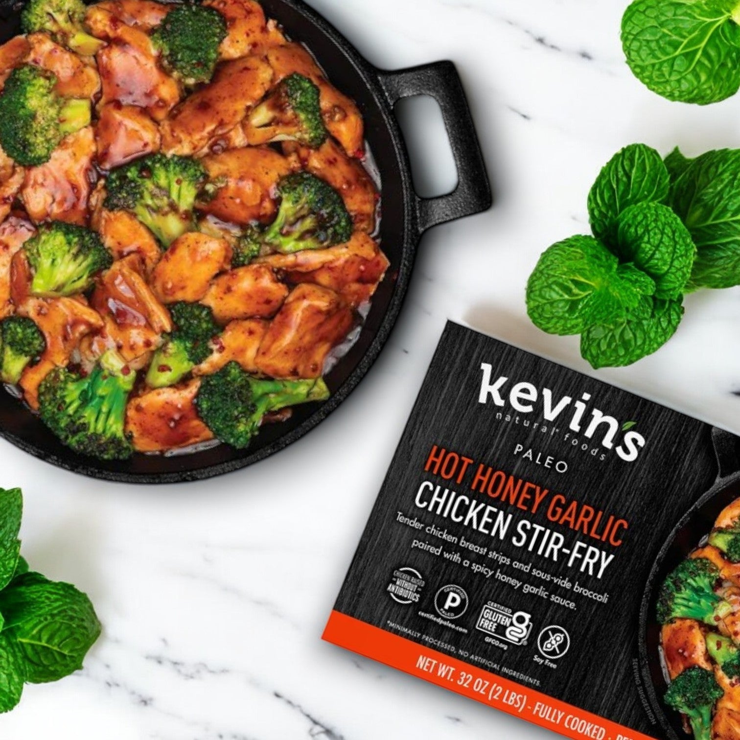 A skillet with antibiotic-free chicken stir-fry and broccoli beside Kevin's Natural Foods Hot Honey Chicken Stir Fry package sits on a marble surface, garnished with mint leaves.