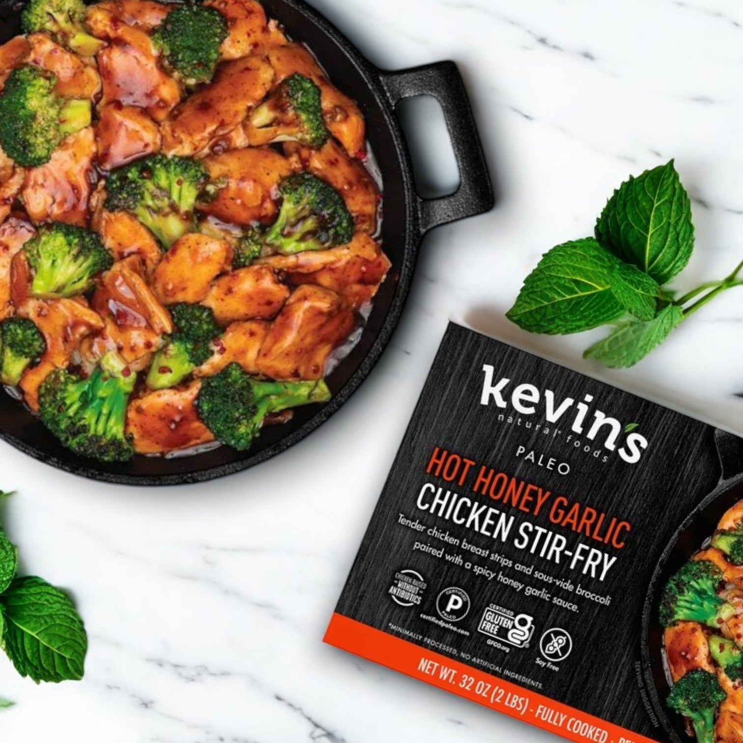 A skillet with Paleo-friendly chicken and broccoli stir-fry sits beside a boxed package labeled "Kevin's Natural Foods Hot Honey Chicken Stir Fry, 32 oz. -3 Count." Garnished with fresh mint leaves, this gluten-free dish from Kevin's is perfect for a quick and healthy meal.