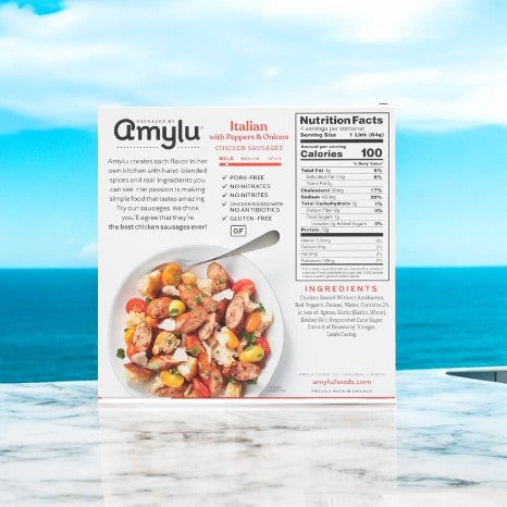 A box of Amylu Italian Chicken Sausage With Peppers & Onions sits on a marble surface by the sea, displaying nutrition facts and ingredients.