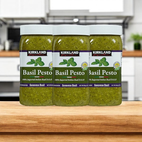 Three jars of Easy Lunches Kirkland Signature Imported Basil Pesto, 22 oz each, sit on a wooden surface against a blurred kitchen backdrop, emphasizing their rich texture and flavor.