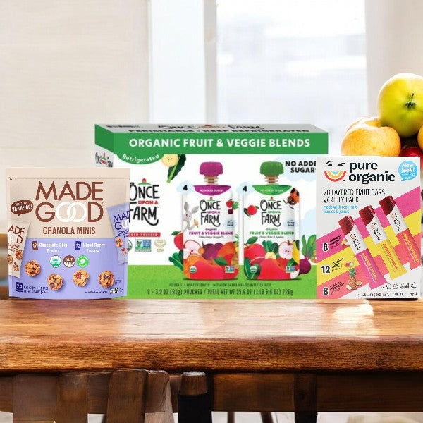 A kitchen counter displaying Easy Lunches Kids Snack Pack! including 24 Made Good Granola Minis, 8 Fruit & Veggie Blends, and 28 Pure Organic Fruit Bars next to a bowl of apples.