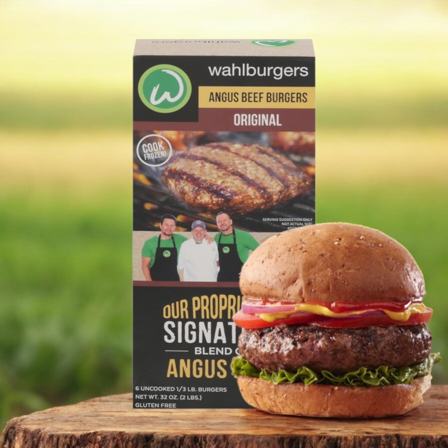 A box of Wahlburgers Angus Beef Burgers, Original, 6 ct, 32 oz sits behind a cooked burger on a wooden surface. Topped with lettuce, tomato, cheese, and ketchup on a bun, it offers restaurant-quality flavor for a premium burger experience.