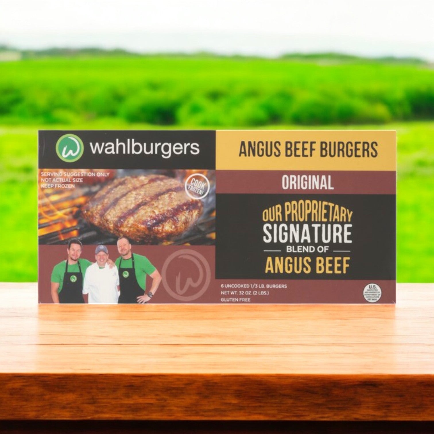 Three boxes of Wahlburgers Angus Beef Burgers, Original, 6 ct, 32 oz are displayed on a wooden surface with images of grilled burgers and chefs, set against a blurred green landscape background.