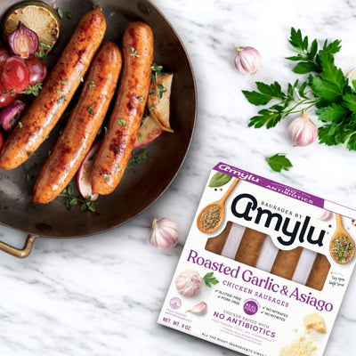 A skillet with roasted sausages and herbs rests beside a pack of Amylu Roasted Garlic & Asiago Chicken Sausage - 4 CT (9 oz) on a marble surface.