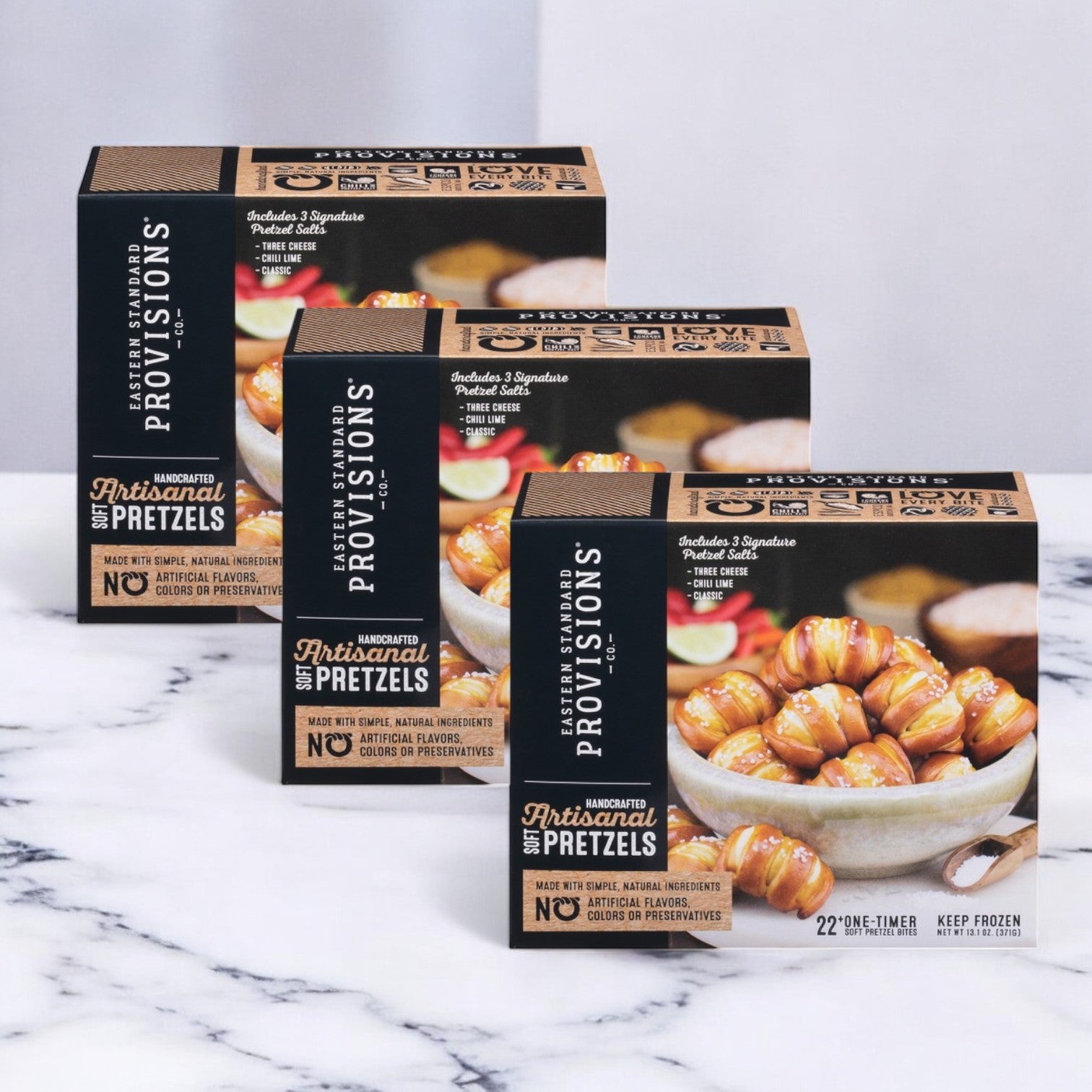 Three packs of 13.1 oz. Eastern Standard Provisions Soft Artisanal Pretzels are stacked on marble, highlighting "No Artificial Flavors, Colors, or Preservatives," reflecting the brand's quality commitment.