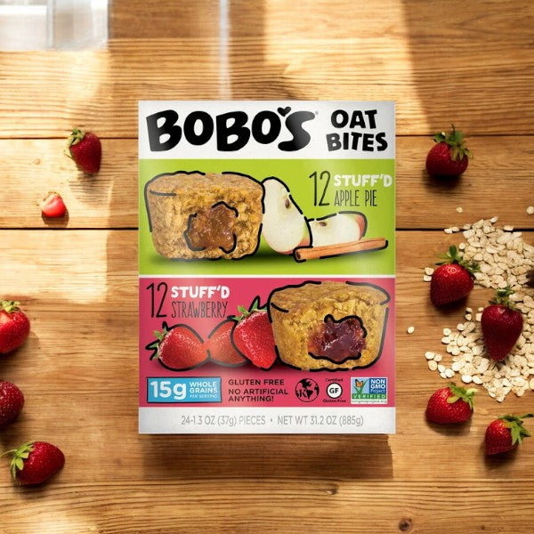 A box of Easy Lunches' Bobos Oat Mini Bites, 1.3 oz - 24 Counts, placed on a wooden surface, surrounded by strawberries and oats. Featuring 12 "Stuff'd Apple Pie" and 12 "Stuff'd Strawberry" flavors, these wholesome treats highlight their whole grain content and come individually wrapped for convenience.