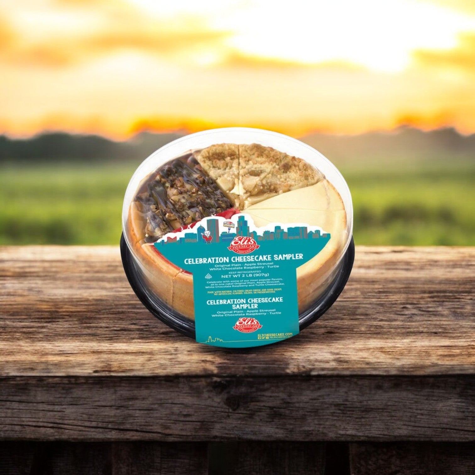 A Connie's Bundle from Easy Lunches, featuring a cheesecake sampler with an array of flavors including classic plain cheesecake, is elegantly presented in a clear plastic container on a wooden surface. The backdrop showcases the warm hues of a sunset over an expansive field. Enjoy free shipping with this bundle!