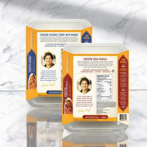 Two packages of Easy Lunches Sukhi Meal Pack! ready-to-eat meals on a marble surface: Chicken Coconut Curry with Mango (left) and Tikka Masala Family Entree Meal (right). Both 15 oz packages display nutritional information and a photo of a woman, offering a taste of vibrant Indian cuisine.