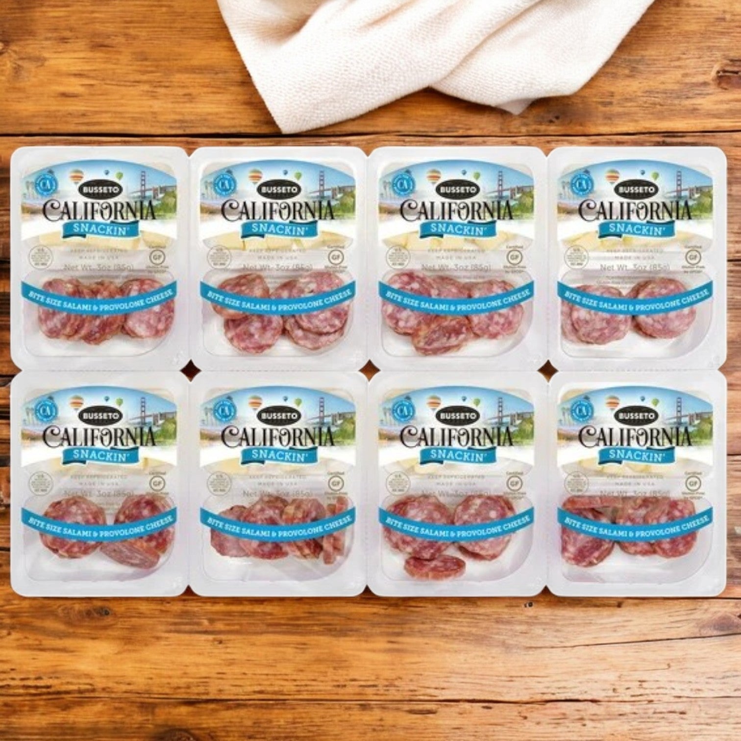 Nine packs of Busseto California Snackin' Bite Size Salami & Provolone Cheese, high in protein, are arranged in a 3x3 grid on a wooden surface with a beige cloth partially visible in the background.