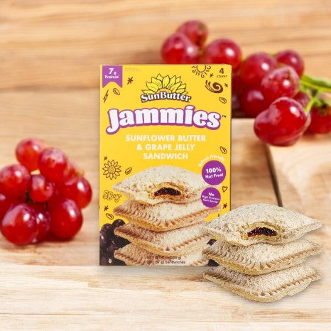 A box of SunButter Jammies Sandwiches, Sunflower Butter & Grape Jelly (1 Box - 4 Sandwiches) from the brand Sunbutter placed on a wooden surface with grapes in the background and two sandwich pieces in the foreground.