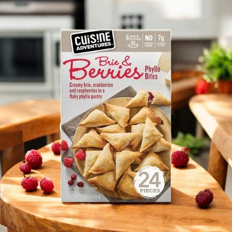 A box of Cuisine Adventures Berries & Brie Phyllo Bites rests on a wooden table, encircled by fresh raspberries. These delicious appetizers boast a flaky phyllo pastry filled with creamy brie and vibrant berries, making them ideal for any gathering.