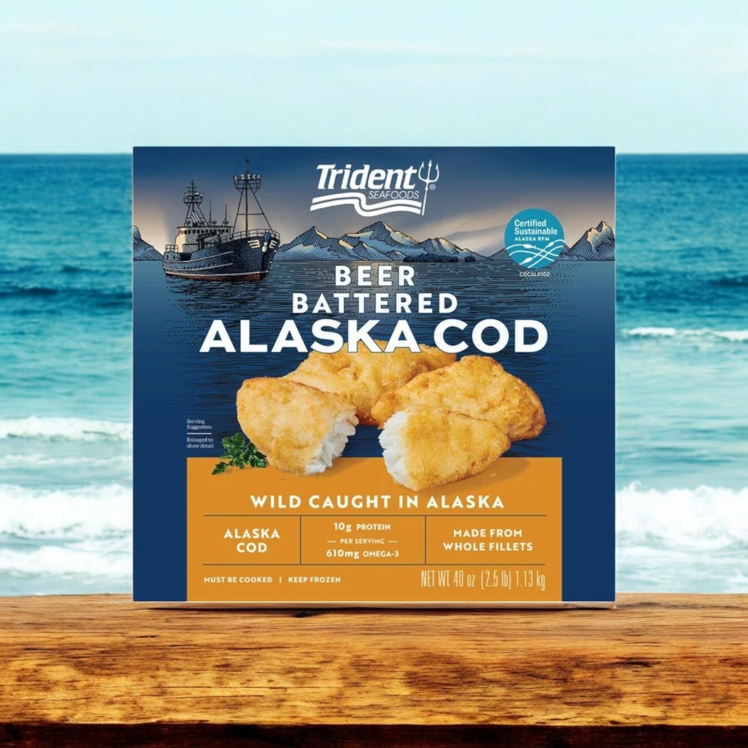 A 2.5 lbs box of Trident Seafoods Wild Alaskan Beer Battered Cod is placed on a wooden table against a seascape backdrop, showcasing its "Wild Caught in Alaska" and "Made from Whole Fillets" attributes that highlight its sustainable, wild origins.