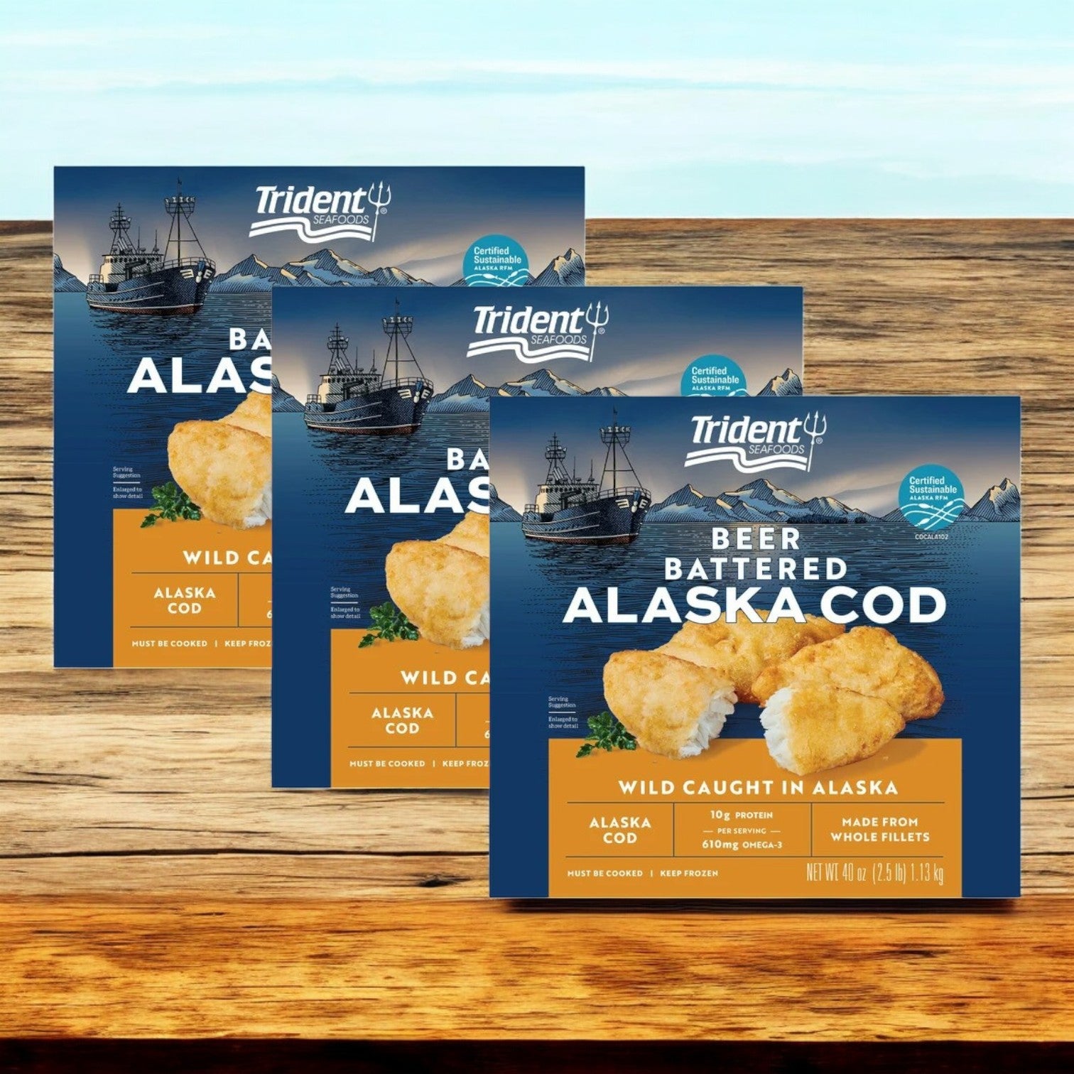 Three packages of Trident Seafoods Wild Alaskan Beer Battered Cod, weighing 7.5 lbs each, rest on a wooden surface with images of fishing boats and a seafood certification logo, promising the delicious taste of sustainably sourced cod with every bite.