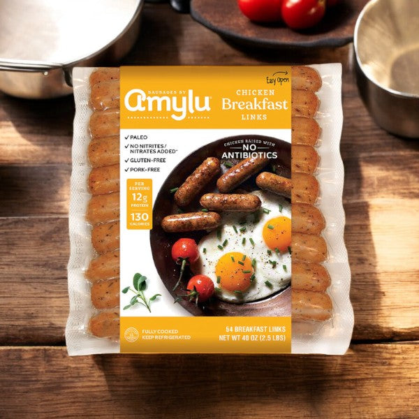 A package of Amylu Chicken Breakfast Sausage Links, weighing 40 ounces and made by the brand Amylu, rests on a wooden table surrounded by a frying pan, tomatoes, and greens. The packaging highlights all-natural ingredients including antibiotic-free choices and paleo options, making it ideal for anyone looking for gluten-free and healthy foods.