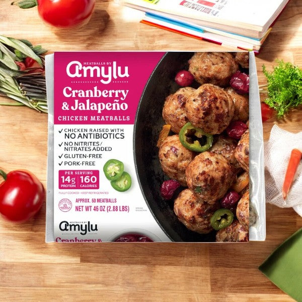 Amylu Chicken Meatballs, Cranberry and Jalapeno, 46 oz, are showcased on a wooden surface surrounded by fresh vegetables. This healthier option features an all-natural ingredient list and is free from antibiotics, gluten, and pork.