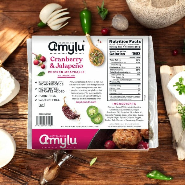 A package of AmyLu Chicken Meatballs, Cranberry and Jalapeno (46 Oz, 1ct), is elegantly displayed on a wooden table decorated with garlic, rosemary, and cranberries. This healthier option by Amylu features all-natural ingredients and conveniently displays its nutrition facts and ingredients directly on the packaging.