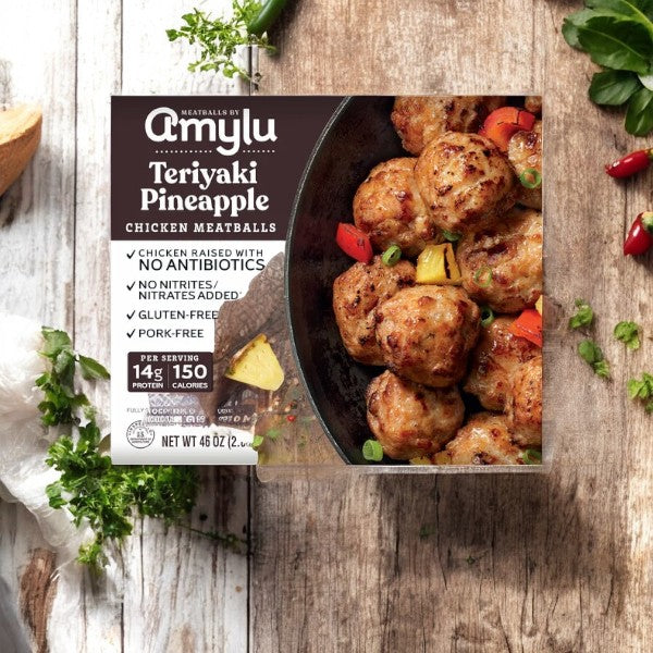 The packaging of Amylu Teriyaki Pineapple Chicken Meatballs, 1ct, 46 oz is displayed on a wooden table with herbs and spices, emphasizing its all-natural ingredients.